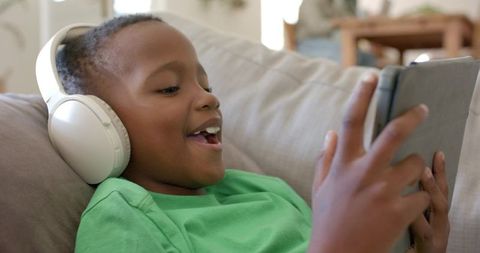Young boy wearing white headphones enjoying tablet on couch, casual streaming and speaking