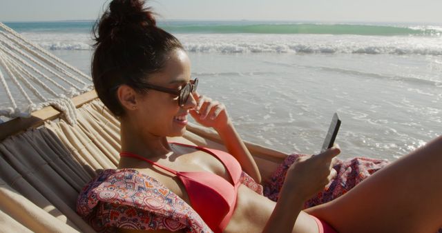 Woman relaxing in a hammock near the ocean, wearing a red swimsuit and sunglasses while using her phone. Ideal for travel blogs, summer vacation advertisements, tropical destination promotions, and leisure lifestyle content.
