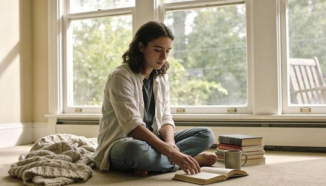 Young man sitting cross-legged reading paperback on carpet near sunny windows with mug