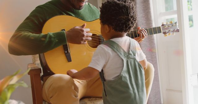 Father playing guitar for young child in a bright sunlit room. Perfect for family bonding concepts, parenting blogs, music education materials, or advertisements focused on nurturing and educational activities between parents and children.