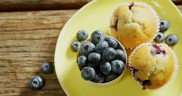 Fresh blueberries accompany delicious muffins on a vibrant yellow plate, with copy space. Blueberries add a burst of flavor and a nutritional boost to baked goods and snacks.
