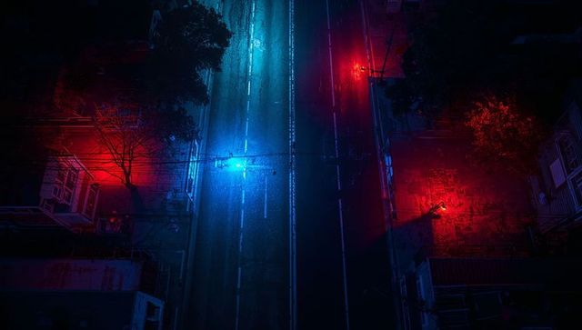 Aerial city street reflecting neon blue glow between red-lit rowhouses at night