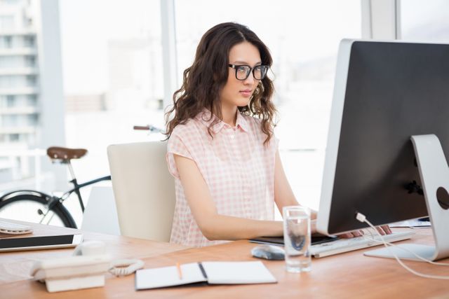 Young hipster woman wearing glasses working on a computer in a modern office. Ideal for use in business, technology, and professional work environment contexts. Can be used for articles on modern workspaces, productivity, and office culture.