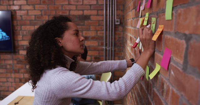 Young woman writing on colorful sticky notes on brick wall in modern office, actively engaging in brainstorming session. Useful for illustrating teamwork, creative processes, project planning, or office dynamics in business environments.