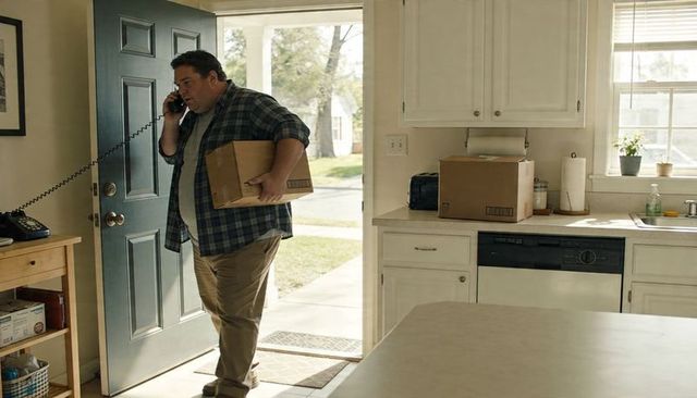 Hispanic man carrying cardboard box talking on corded landline while entering kitchen