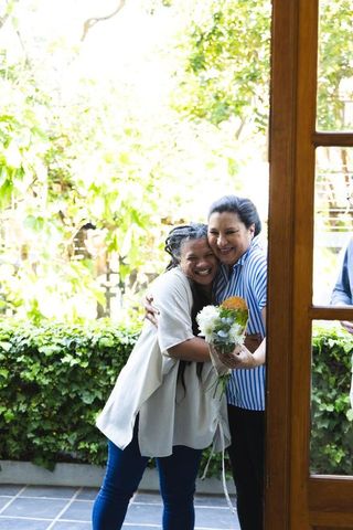 Joyful friends hugging on doorstep holding small orange and white bouquet, candid reunion