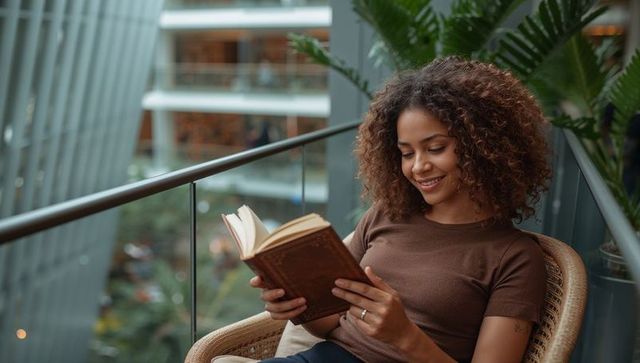 Smiling woman reading hardcover book on wicker balcony chair in modern glass atrium