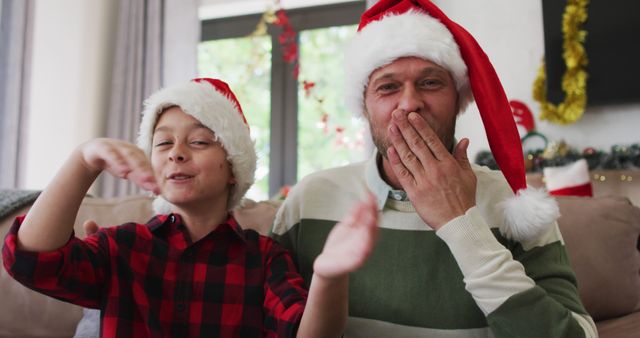 Father and son are sitting on a cozy couch at home, both wearing Santa hats. They are enjoying Christmas and bonding together. Background features festive Christmas decorations, contributing to the warm and joyful holiday atmosphere. This image is perfect for advertisements, holiday greeting cards, family-oriented marketing campaigns, and festive social media posts.