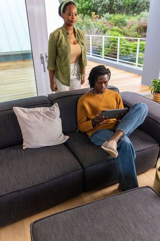 African american couple sitting on sectional sofa using tablet in sunlit modern living room