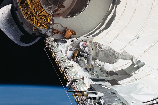 Astronaut James H. Newman, mission specialist, uses a 35mm camera to take a picture of fellow astronaut Carl E. Walz (out of frame) in Discovery's cargo bay. The two were engaged in an extravehicular activity (EVA) to test equipment to be used on future EVA's. Newman is tethered to the starboard side, with the orbital maneuvering system (OMS) pod just behind him.
