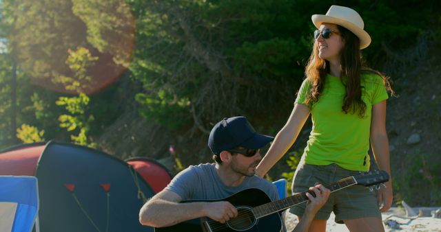 Young man plays guitar while woman in hat and sunglasses stands next to him, smiling. They are at an outdoor campsite with tents and trees in background. Ideal for use in articles or advertisements related to summer activities, music, camping, adventure, and bonding experiences in nature.