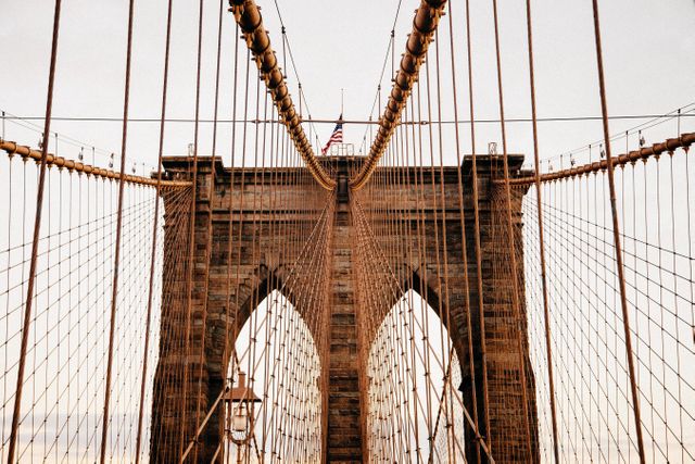 The iconic stone towers and symmetrical cables of the Brooklyn Bridge stand prominently with an American flag visible at the top, captured at dusk. This image can be used for travel and tourism content, emphasizing architectural highlights, or illustrating iconic American landmarks. Suitable for websites, magazines, posters, and promotional material highlighting New York City or classic American architecture.