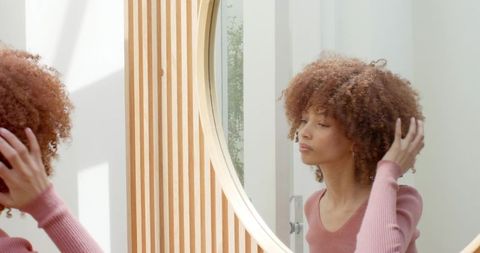 Young woman adjusting hoop earring at round mirror with natural light and wood slats