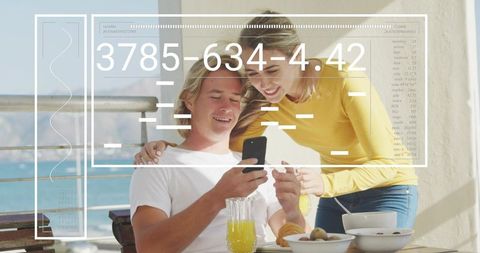 Couple sharing sunny oceanfront breakfast on balcony while checking smartphone with hud