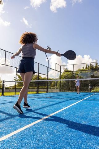 Diverse women playing padel on blue court jumping for volley under sunny sky