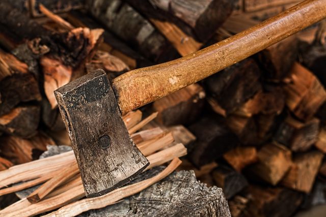 Detailed close-up of an axe embedded in a piece of chopped wood with a pile of logs in the background. Depicts traditional wood splitting in a rustic outdoor environment. Suitable for content related to manual labor, sustainable practices, camping, survival skills, or imagery representing strength and endurance.