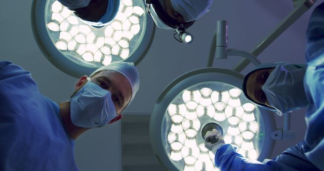 Surgeons in operating room are focusing on patient under bright surgical lights. They are wearing surgical masks and gowns, emphasizing a sterile environment. Use this for healthcare promotions, medical articles, illustrating medical procedures or hospital operations, and medical teamwork scenarios.
