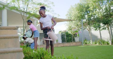 Mom and daughters watering garden plants with watering cans on sunny backyard lawn