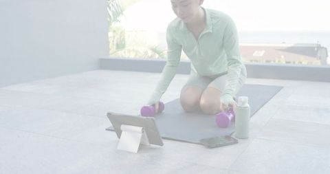 Midadult woman kneeling on balcony following online workout with purple dumbbells in mint zipup top