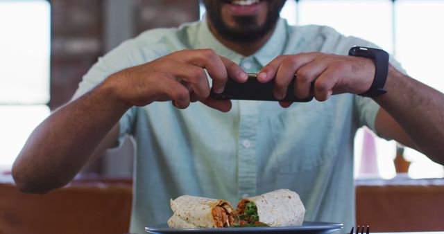 Man enthusiastically capturing photo of burrito with smartphone in casual dining environment. Perfect for use in food blogs, social media marketing, culinary websites, restaurant promotions, or visual content related to food enthusiasts.