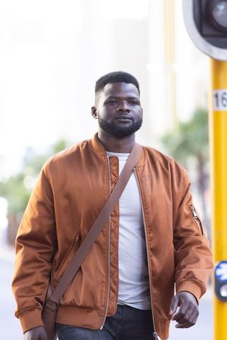 African american man walking city street wearing rust bomber jacket and crossbody bag