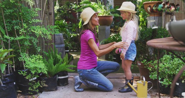 A mother and her young daughter are spending quality time together in a vibrant garden or plant nursery. The mother is kneeling down and talking to her daughter, both wearing hats and casual clothes. Suitable for use in articles about family activities, parenting, gardening hobbies, and healthy outdoor living.