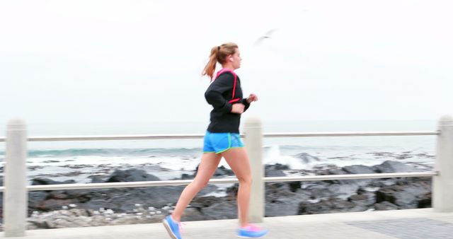 A young Caucasian woman is jogging along a seaside promenade, with copy space. Her active lifestyle is emphasized by the dynamic backdrop of the ocean and open sky.
