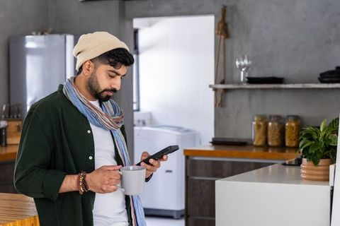 Young man in beanie and scarf checking smartphone while holding mug in modern kitchen