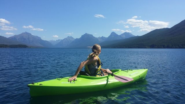 Woman enjoying kayaking on a calm mountain lake with clear skies and distant peaks. Perfect for articles or promotions about outdoor adventure, travel destinations, summer activities, and nature exploration.