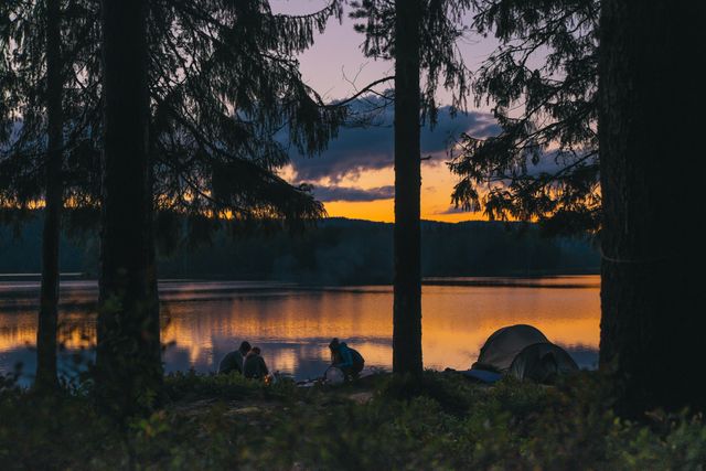 Group camping beside a calm lake during sunset. A picturesque forest scene with tents and silhouettes reflected on the water, suggesting adventure, relaxation, and a connection with nature. Ideal for travel blogs, outdoor adventure promotions, nature retreats, and camping equipment advertisements.