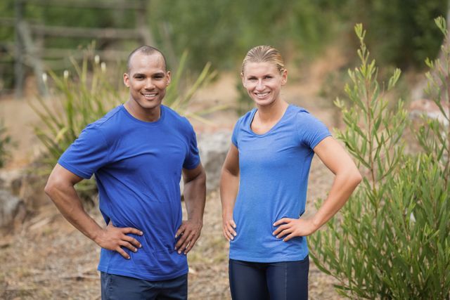 Fit man and woman standing with hands on hips during outdoor boot camp training. Both are wearing blue sportswear and smiling confidently. Ideal for use in fitness, health, and lifestyle promotions, as well as advertisements for outdoor training programs and athletic gear.