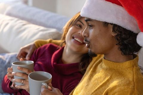 Couple cuddling on sofa wearing santa hat and knit sweaters holding ribbed mugs of hot drink