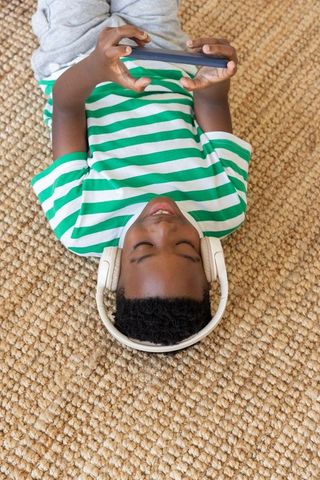 African american boy lying on woven rug wearing headphones and holding smartphone