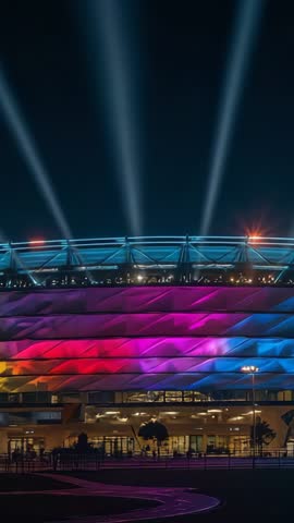Arena facade lighting up in cycling LED colors with searchlights beaming at night vertical video