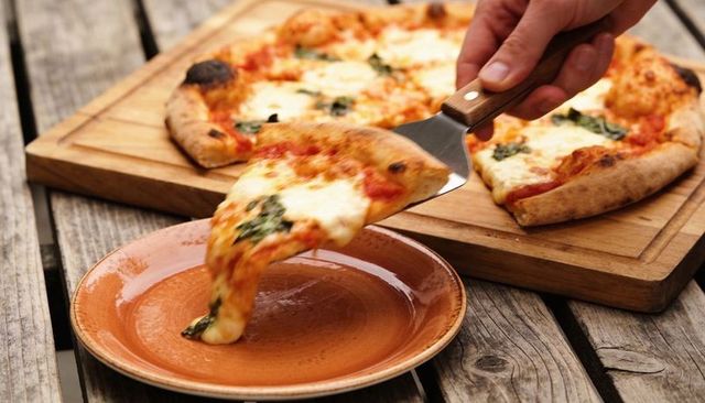 Hand lifting wood-fired margherita pizza slice with stretchy mozzarella over clay plate