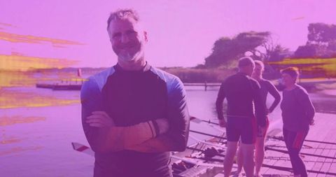 Confident mature rower standing with arms crossed on dock beside rowing shells and crew