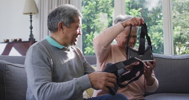 Elderly couple engaging with virtual reality headsets in a cozy living room. The couple is smiling and appearing curious, indicating enjoyment and novelty. Ideal for promoting technology products designed for seniors, senior lifestyle content, retirement home brochures, and articles showcasing the incorporation of modern technology into older adults' lives.