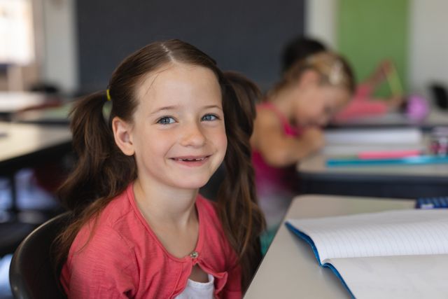 Young girl with pigtails smiling while sitting at her desk in a classroom. Ideal for educational materials, school advertisements, and back-to-school promotions.