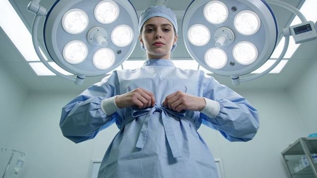 Female surgeon tying surgical gown under operating lights preparing for surgery sterile or