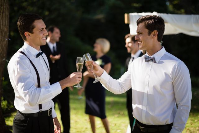 Waiters in formal attire are toasting with champagne glasses at an outdoor wedding in a park. They are smiling and appear to be enjoying the celebration. The background shows other guests and greenery, indicating a festive and joyful atmosphere. This image can be used for wedding planning, event management, hospitality services, and celebratory occasions.