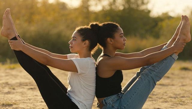 Sunlit partner yoga v pose back-to-back young women practicing outdoor flexibility and balance