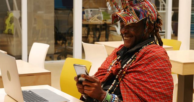 Man in traditional African attire smiling while using a smartphone and sitting near a laptop in a modern workspace. Can be used for cultural diversity, technology in diverse settings, or blending tradition with modern lifestyle themes.