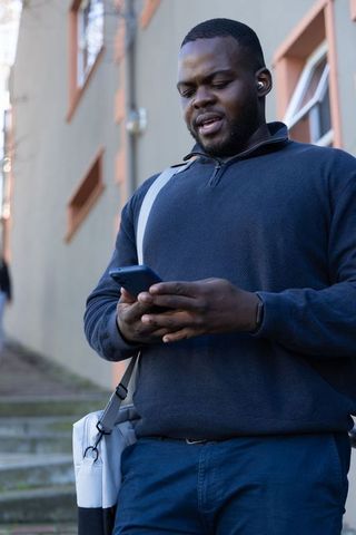 African american commuter checking smartphone while walking with messenger bag and earbuds