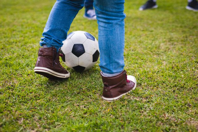 Low section of a child playing football on a grassy field. The child is wearing jeans and brown sneakers, engaging in an outdoor activity. Ideal for use in articles or advertisements related to children's sports, outdoor activities, healthy lifestyles, and childhood hobbies.