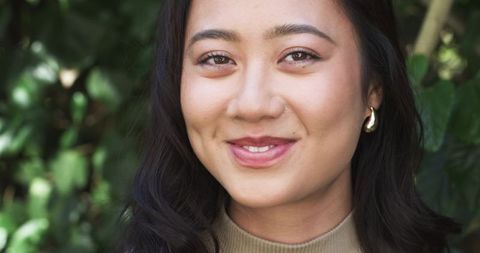 Smiling asian woman posing in garden wearing beige sweater and gold hoop earring