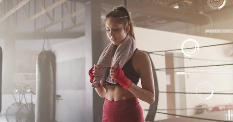 Young woman boxer pausing with towel and wrapped hands beside punching bag in gym