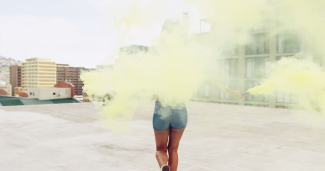 Young biracial woman enjoys a vibrant smoke bomb outdoors. She creates a playful atmosphere on a rooftop with a colorful display.