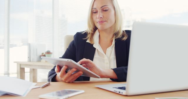 Blond businesswoman using tablet in modern office, laptop and papers on desk, reflecting advancement in digital technology at work. Ideal for articles or ads related to professional environments, technology integration, women's empowerment in business, remote work solutions.