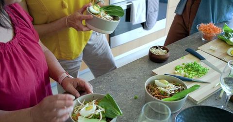 Diverse women including older woman preparing fresh noodle bowls at kitchen island together