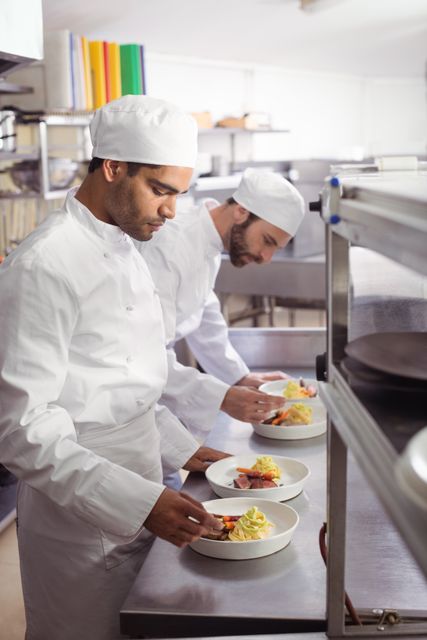 Two chefs dressed in white uniforms are carefully garnishing dishes with a professional touch in a commercial kitchen. Perfect for marketing materials, culinary arts publications, restaurant advertisements, or hospitality training programs.